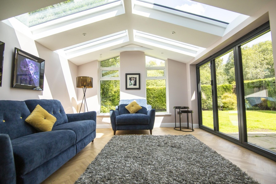 A well-lit living room with a navy blue sofa and armchair. The room features a vaulted ceiling with two large Hup! skylights and multiple windows offering a view of a green garden. The floor is a light wood in a herringbone pattern, partially covered by a shaggy gray rug.